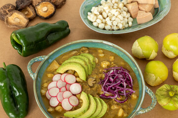 Vegan Pozole verde topped with sliced avocado, radish and red cabbage showed with raw main ingredients: hominy,poblano pepper, fresh tomatillo, young jackfruit and dried shiitake mushrooms.