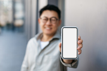 Asian businessman demonstrating blank smartphone screen at camera, recommending new mobile app or website, mockup