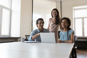 Fototapeta premium Happy successful business team posing at shared workplace, looking at camera, smiling, laughing. Group working at laptop computer office portrait. Teamwork, communication concept. Low angle