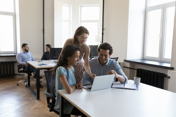 Diverse group of cheerful employees sharing computer at office table, talking, laughing together. Corporate mentor training interns, showing online presentation, work app on laptop, checking task