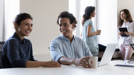 Happy cheerful diverse coworkers chatting at workplace, laughing, enjoying teamwork. Two employees...