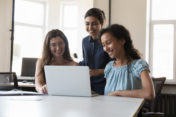 Happy successful diverse team of three employees women using laptop together at shared workplace, looking at monitor, laughing, talking on video conference call, enjoying online communication