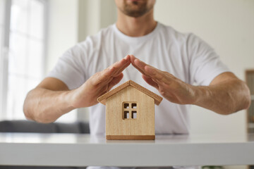 Man makes roof protection over small wooden house with his hands, symbolizing property and security insurance. Close up of house model on table and man's hands above it. Home insurance concept.