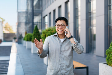 Happy successful asian businessman talking on smartphone, standing outdoors against modern office...