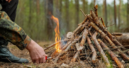 Man starts a fire in the forest using a lighter. Close-up of a man's hand lighting a fire with a lighter. Lighting a fire in the forest by a person. Low angle of a burning fire made of brushwood... - obrazy, fototapety, plakaty