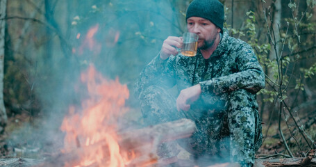 Fototapeta premium Man with cup of tea the fire in the forest. Human looks at the bonfire and drinks tea on the nature in the summer. Alone traveler sits and rests by the fire with a cup of coffee at dusk.