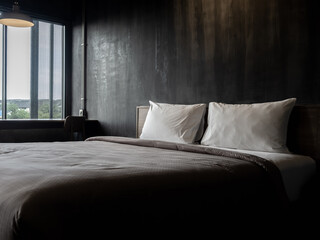 Couple white pillow on the empty bed with brown duvet on dark grunge concrete wall background near the window in hotel bedroom loft style. Two soft and clean comfort pillows preparing for the guest.