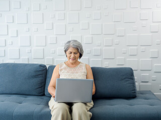 Portrait of smiling Asian senior casual woman sitting on the sofa, working with laptop computer in bright white room. Elderly female surf internet or video call at home. Older people with technology.