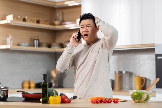 Shocked asian man cooking meal and talking on phone