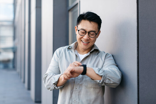 Cheerful asian businessman wearing smart watch and looking at it, checking time, using modern gadget outdoors