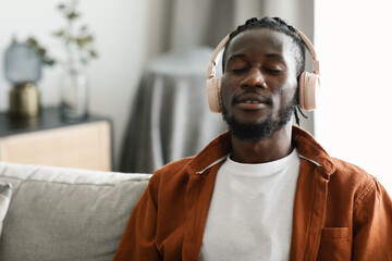 Calm african american man in wireless headphones sitting on sofa with closed eyes and listening to...