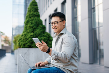 Mature asian man with laptop sitting outdoors and texting on smartphone, sitting outdoors near office building