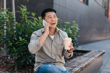 Happy asian male entrepreneur talking on smartphone and drinking coffee from takeaway cup, sitting outdoors