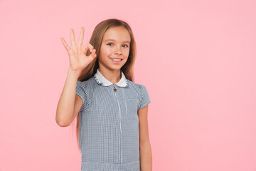 Young smart caucasian preteen teenager girl schoolgirl daughter in blue dress looking at camera showing okay gesture isolated in pink background - Powered by Adobe