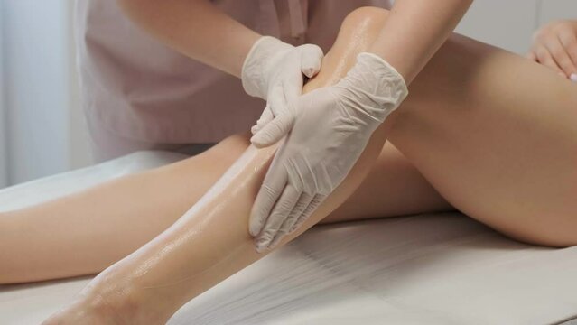 A female depilatory expert applies sugar paste on the patient's leg. The process of hair depilation on the girl's leg with sugar paste by a professional master.