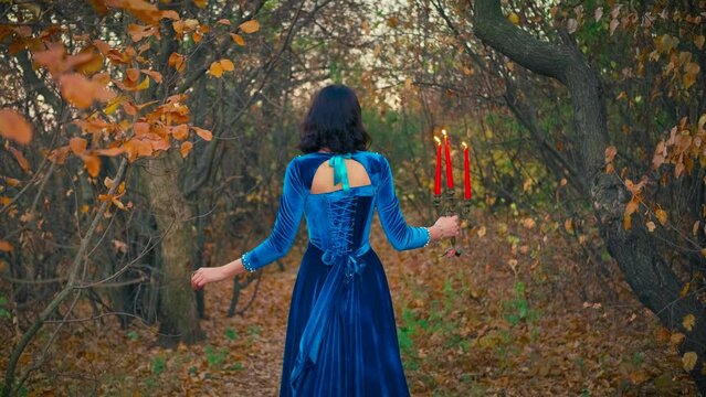 Beauty autumn forest, yellow leaves, bare black tree trunks. Silhouette elegant fantasy woman. Brunette lady turned away, holding candlestick, red candles burning bright fire. Long blue vintage dress