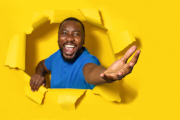 Overjoyed young black man stretching open palm through hole in torn yellow paper background, taking...
