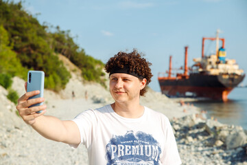 smiling Young Caucasian man with curly hair making selfie with smartphone. Guy outdoors against sunken cargo ship and taking self portrait with mobile phone. Summer head shot lifestyle portrait