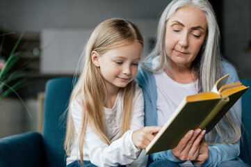 Serious caucasian small granddaughter and mature grandmother are reading book on sofa in room interior