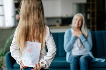 European small granddaughter holds behind back postcard with picture of happy senior female