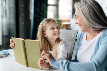 Cheerful caucasian little preschooler with aged grandma read book, enjoy study and free time in living room