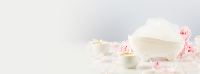 Wellness spa banner with mini bathtub with soap foam, candles and rose petals on a white table with copy space. Concept of bath time, self care and relaxing time. Soft focus style image, Banner size