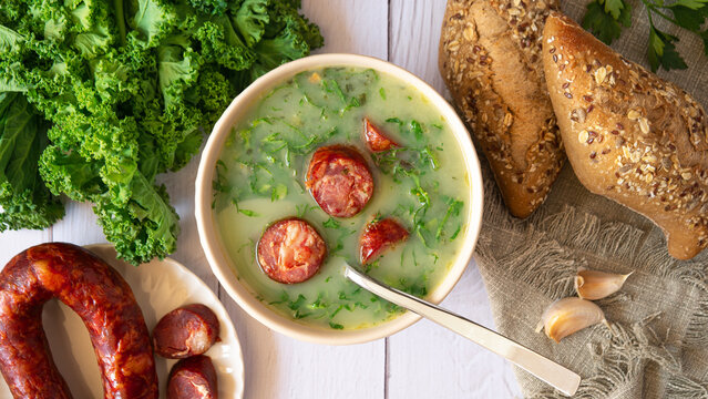 Portuguese style soup called Caldo Verde, bread, Cabbage julienne, and chorizo sausage on a white wooden Table. Flat lay