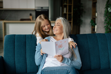 Glad pretty caucasian small granddaughter hugs retired grandmother gives postcard with drawing