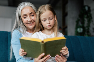 Happy caucasian little granddaughter and senior lady reading book on sofa in living room interior