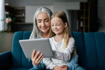 Satisfied caucasian small girl and old lady watch video, enjoy online game on tablet, have call on...