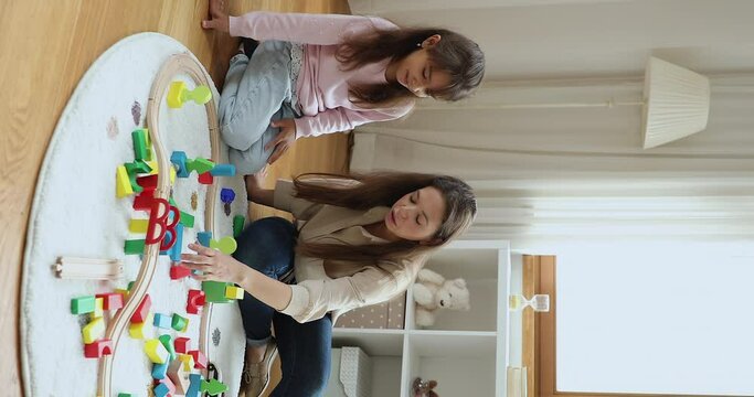 Happy active young mother and daughter kid constructing toy city road, highway model on heating carpeted floor, arranging, stacking colorful building blocks, talking, playing game at home