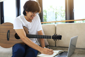An Asian man in a white shirt writes down ideas from his acoustic guitar in a notebook as a weekend...