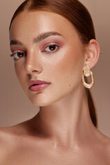 Female model with red hair, brown eyes, freckled skin, poses for a beauty shoot. She wears gold...