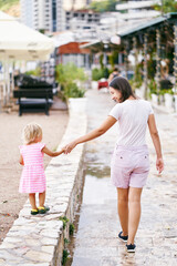 Smiling mom leads a little girl by the hand along a stone high curb on the street. High quality photo