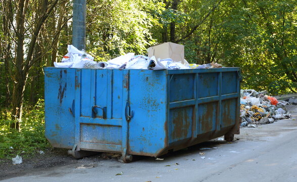 A large blue metal dumpster filled with garbage stands near the road