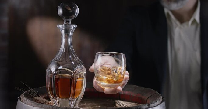 Man in a suit and beard (sommelier) takes a glass of whiskey on ice from a vintage whiskey barrel and shakes the glass to spread the smell of whiskey. Slow motion 4K