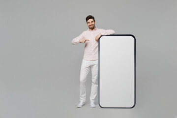 Full body young caucasian man he wear basic white shirt point finger on big huge blank screen mobile cell phone with workspace copy space mockup area isolated on plain grey background studio portrait.