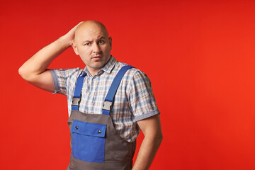 Bald man in work clothes looks at the camera in amazement and holds the back of his head with one...