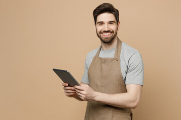 Young smiling man barista barman employee in brown apron work wear coffee shop hold use tablet pc computer look camera isolated on plain pastel light beige background. Small business startup concept.