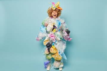 Full length shot of emotional female activist engaged in antipollution campaign poses in costume made of garbage and litter on head shouts loudly isolated on blue wall. Save planet from contamination