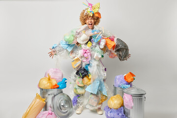 Indoor shot of emotional woman screams loudly made costume from plastic garbage keeps mouth widely opened demands to stop destroying our planet poses near bins full of litter. Environmental protection