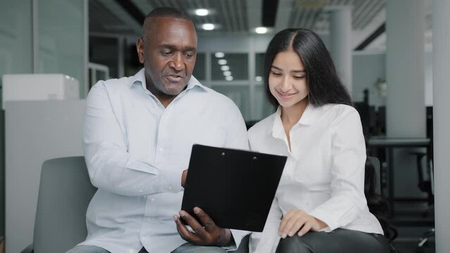 Diverse business partners sit in office discuss paper instruction african businessman show documentation in folder tablet to arabian female colleague explain insurance contract benefits negotiating