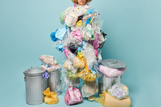 Cropped shot of unknown person poses plastic garbage costume surrounded by dustbins being aware of environmental pollution and ecological problem isolated over blue background. Say no to contamination