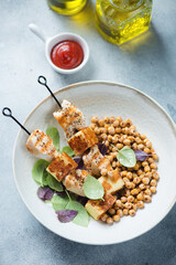 Skewers with roasted chicken and halloumi, fried chickpeas and basil served in a beige plate, elevated view on a light-blue stone background, vertical shot