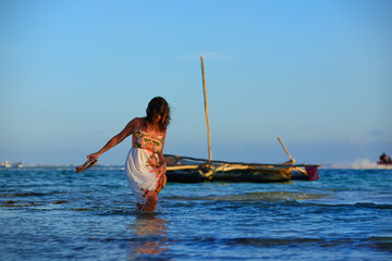 A dark-haired woman in a colourful dress wading through the clear sea, with a native dhow boat and blue sky in the background. Zanzibar, holiday on the Indian Ocean beach. 
