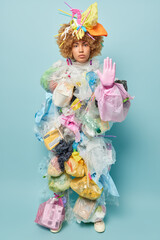 Serious confident woman keeps palm raised towards camera makes forbidden gesture demands to stop polluting environment realizes seriousness of big environmental problem stands against blue wall