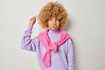 Photo of pensive millennial girl curls hair pouts lips looks thoughtfully aside considers something contemplates about future life dressed in casual purple jumper stands indoor. Let me think
