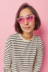 Lovely young dark haired Asian woman looks away thoughtfully daydreams about something wears trendy sunglasses and casual striped jumper isolated over pink background. People and thoughts concept