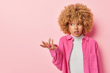 Horizontal shot of confused woman shrugs shoulders has questioned puzzled expression purses lips dressed in shirt isolated over pink background empty space for your advertising content. So what