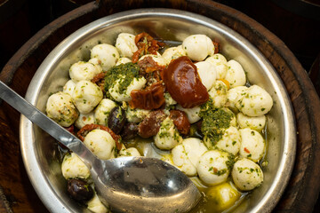 A large mixed vat of mozzarella cheese, olives, and sundried tomatoes in a silver bowl stored in bulk ready to sell smaller measures to consumers in a market looking for fresh home made produce 